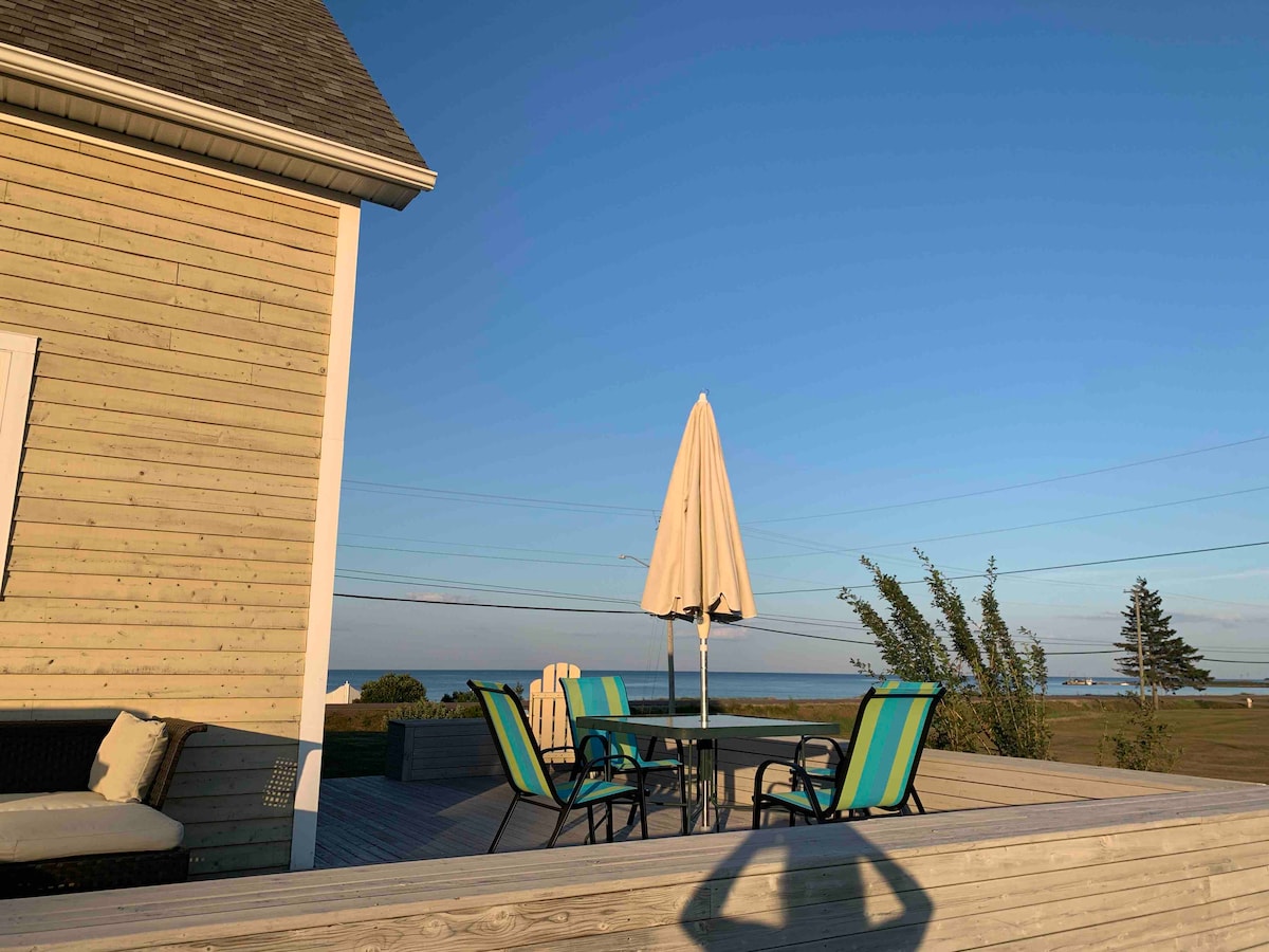 A spacious wooden deck is featured, equipped with a dining table and four colorful chairs. A large umbrella is positioned above the table, providing shade. In the background, the expansive view of the ocean and clear blue sky can be seen.