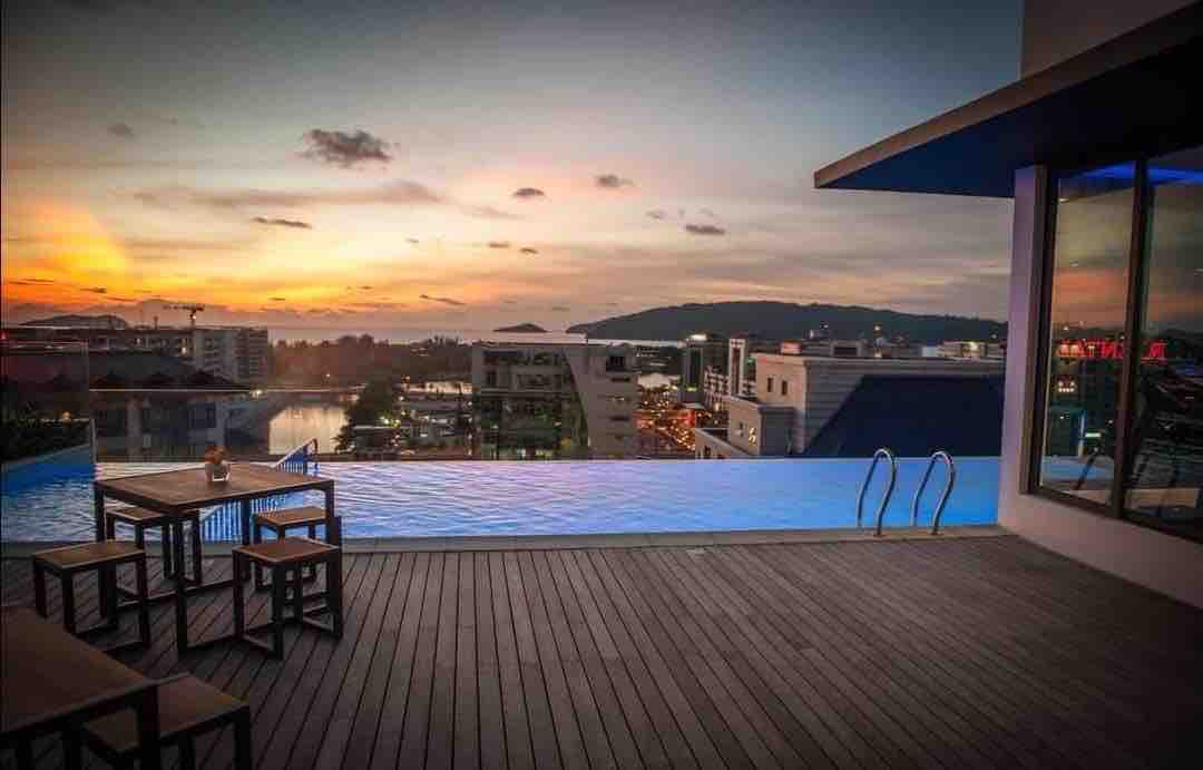 An infinity swimming pool is positioned against a stunning sunset view, offering a panorama of the city skyline. Wooden decking frames the pool area, which includes a set of outdoor dining furniture at one side, providing a serene setting for relaxation.