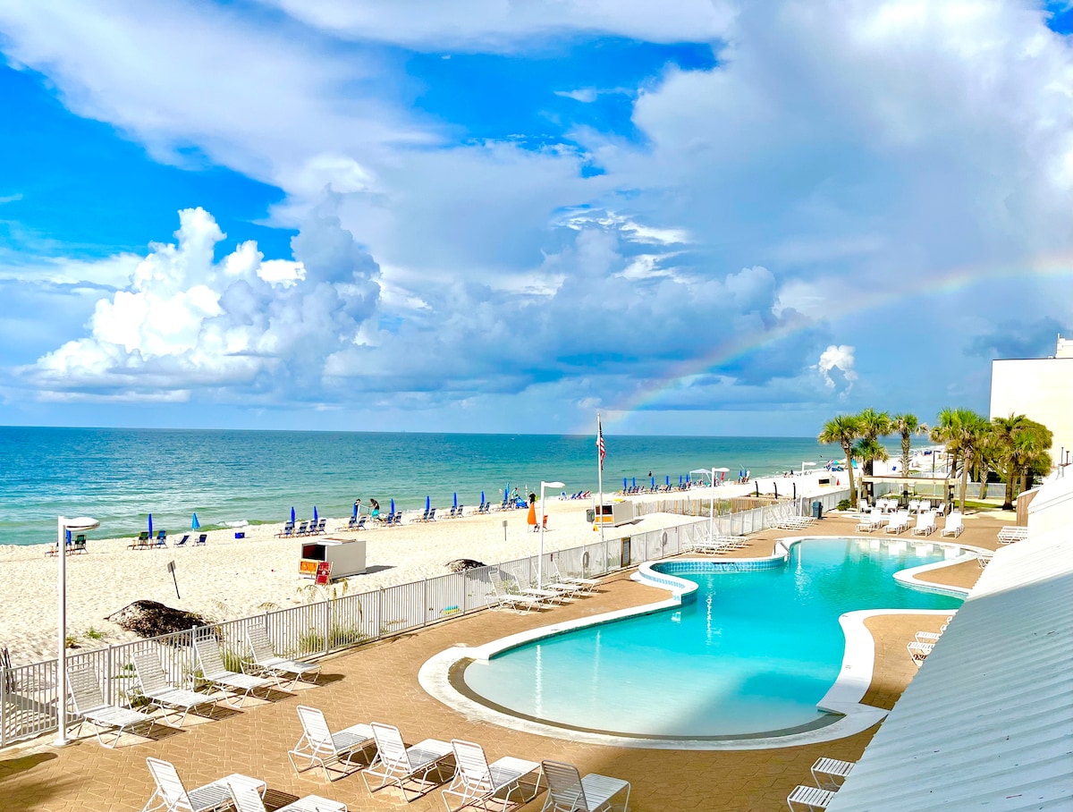 A view of the beachfront property shows a curvy swimming pool, surrounded by lounge chairs, leading to a sandy beach. The ocean sparkles under a blue sky, with wispy clouds and a faint rainbow arching in the distance.