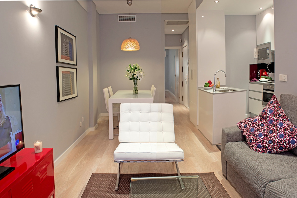 The open-plan living area features a contemporary sofa and a stylish lounge chair. A dining table set for four is visible, along with a well-equipped kitchen in the background. Bright lighting enhances the modern design, complemented by framed artwork on the walls.