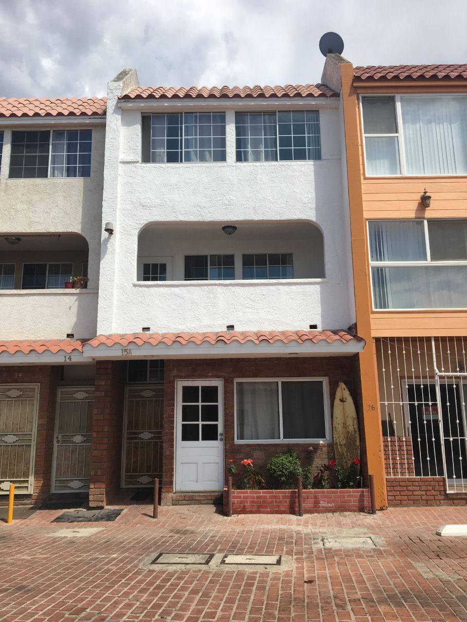 A three-story building features a white exterior with a terracotta tiled roof. The front entrance showcases a simple door framed by a brick facade, with potted plants present in a small garden. Large windows provide ample natural light, complemented by a surfboard displayed nearby.