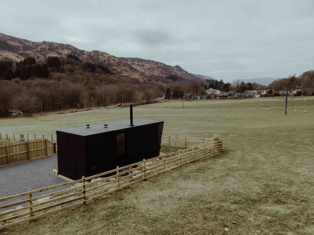 A modern black cabin is set against a backdrop of rolling hills, surrounded by open green fields and a wooden fence. The scenery includes distant trees and a sparse village visible on the horizon under an overcast sky.