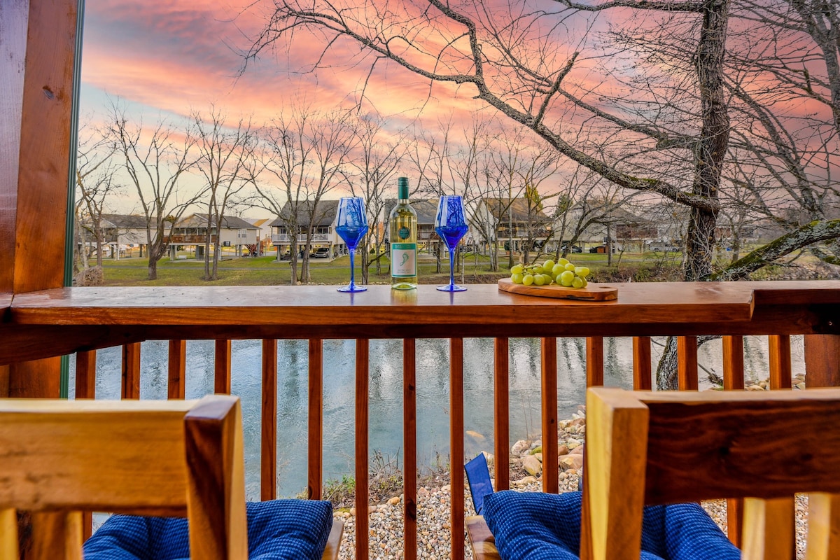 A wooden balcony railing frames a serene view of the river at sunset. A bottle of wine and two blue glasses sit on a table next to a wooden platter of grapes, highlighting a tranquil outdoor setting.