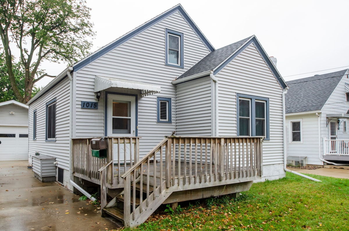 The exterior of the home is characterized by light gray siding and a welcoming front deck with railings. Large windows allow natural light to illuminate the space. The yard features green grass, providing an inviting atmosphere. A driveway and garage are visible in the background.