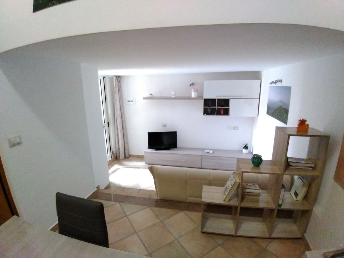 A minimalist living area is depicted, featuring a low wooden TV stand with a flat-screen television. Light-colored furniture is complemented by neutral walls. Shelves display decorative items, while a window allows natural light to fill the space, creating a bright and inviting ambiance.