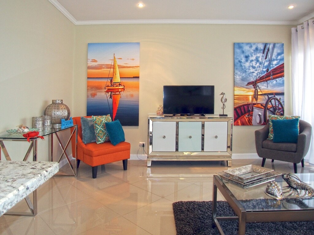 A modern living area features two large artworks depicting serene water scenes, accompanied by a sleek television and stylish furniture. An orange accent chair adds a pop of color, while a glass-topped coffee table sits atop a plush area rug.