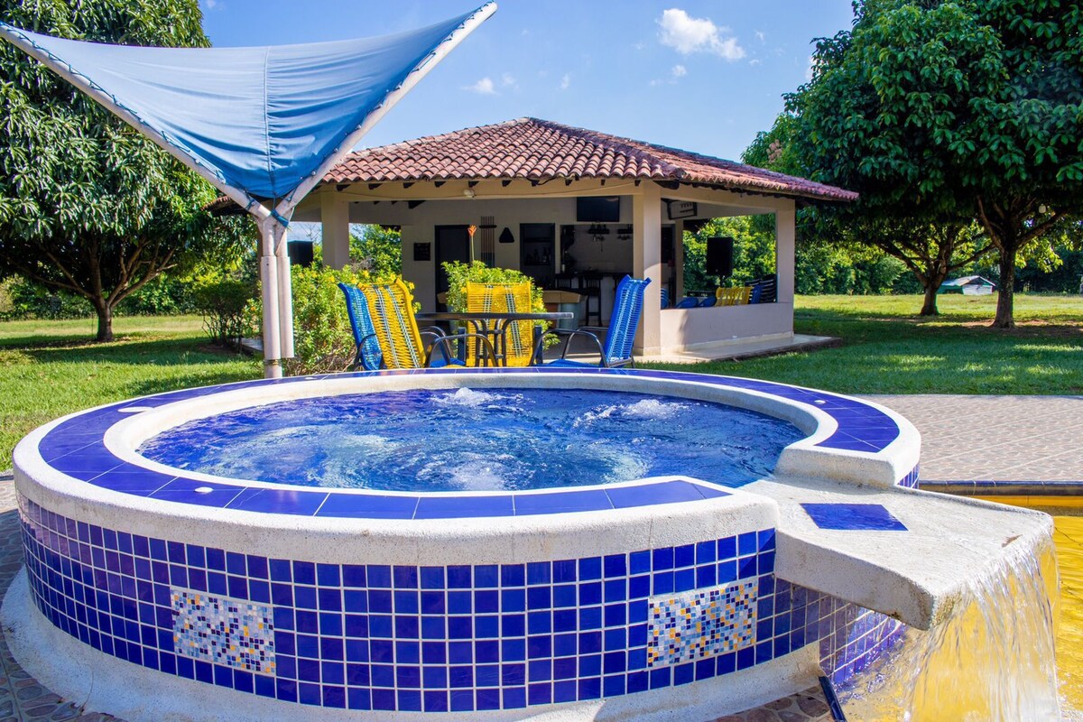 A round jacuzzi is set against a backdrop of green trees and a covered seating area, featuring a table and chairs in the distance. The jacuzzi is surrounded by blue mosaic tiles and has a gentle waterfall feature, inviting relaxation in a natural environment.