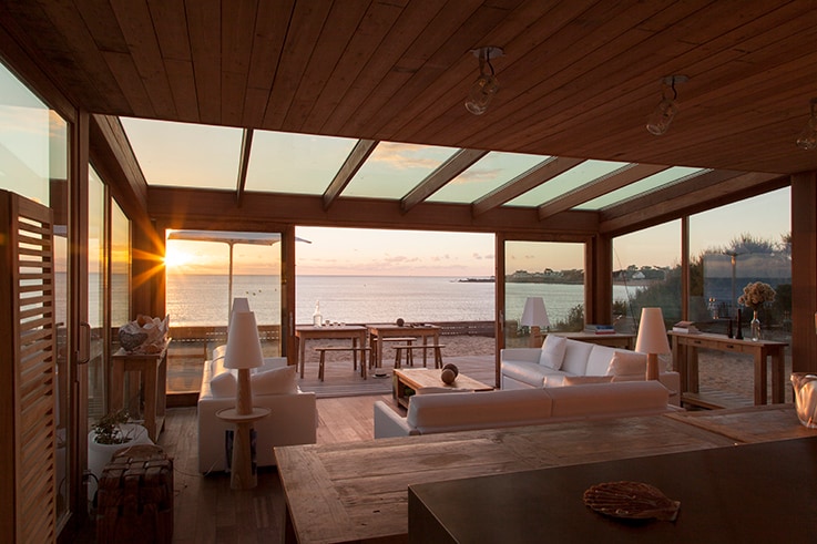 A spacious living area is surrounded by large glass panels, offering an unobstructed view of the ocean and sunset. Comfortable seating is arranged around a wooden table, while natural light fills the room, highlighting the warm wood tones of the ceiling and flooring.