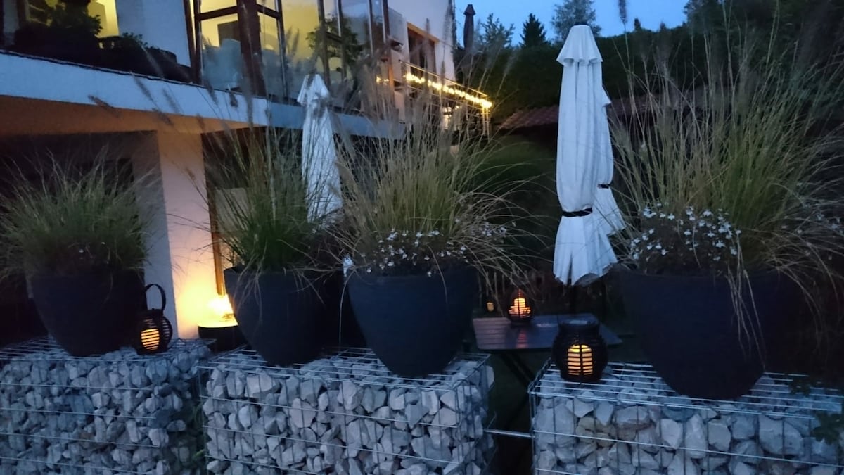 Outdoor seating is framed by tall planters filled with ornamental grasses and small white flowers. Soft lighting creates a calming ambiance, while a table and chairs can be seen in the background, along with umbrellas for shade.