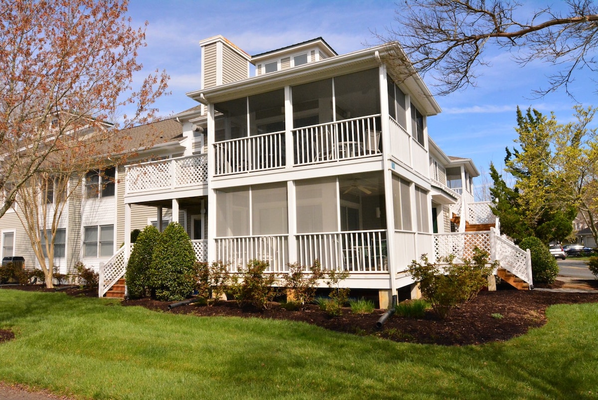 The exterior of a two-story condo is showcased, featuring a large wraparound screened porch. Lush green grass and well-maintained shrubs surround the building, which is complemented by a clear blue sky. Stairs lead up to the entrance, enhancing accessibility.