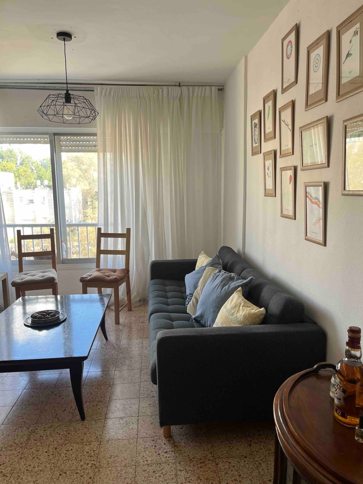 A living area features a dark sofa adorned with decorative cushions, complemented by a wooden coffee table. Two wooden chairs are positioned nearby. Framed artwork is displayed on the wall, and a large window allows natural light to filter into the space, with sheer curtains partially drawn.