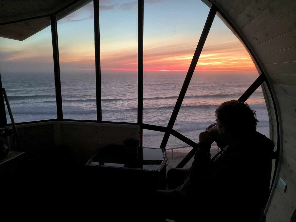 A large window frames a stunning view of the ocean at sunset, where vibrant colors blend seamlessly into the horizon. A person is seated in profile, gazing thoughtfully out at the ocean waves, with a small table placed beside them.