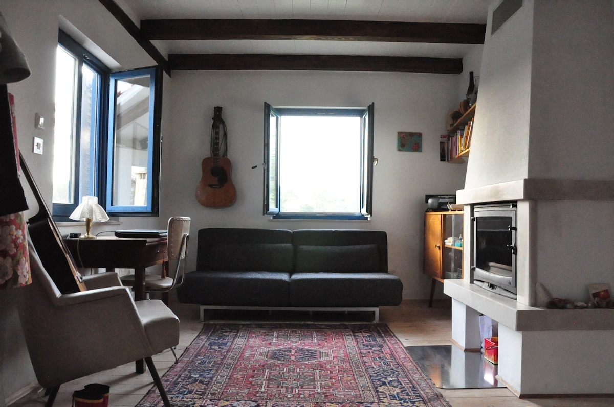 A cozy living area features a dark sofa facing a fireplace, with a vintage rug beneath. Large windows framed in blue allow natural light to enter. A wooden table and a comfortable chair are positioned beside a music instrument, enhancing the room's artistic vibe.