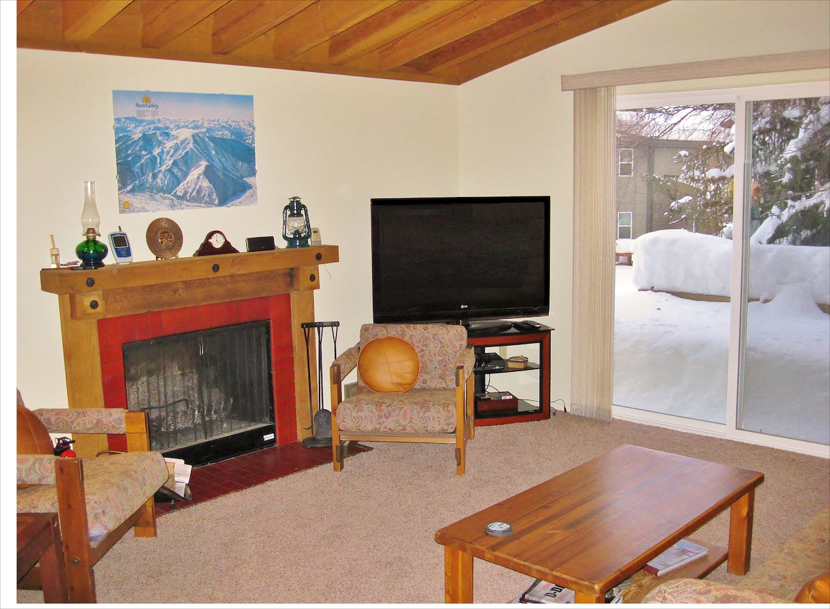 A cozy living area is shown, featuring a wooden beam ceiling and a stone-framed fireplace. Two patterned chairs flank a wooden coffee table, and a large flat-screen TV is positioned against the wall. A sliding door reveals a snowy outdoor scene.