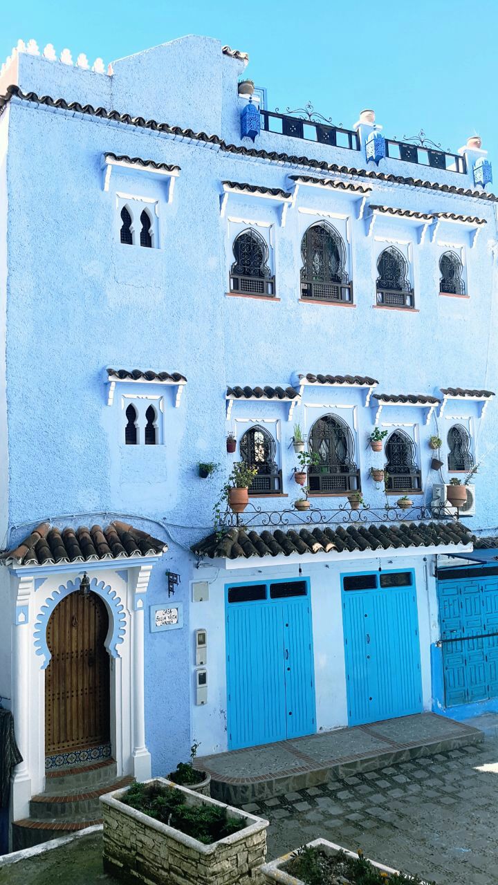 A traditional blue house is displayed with ornate windows and balconies adorned with potted plants. The entrance features an intricately designed wooden door, and two blue garage doors are visible below. The vibrant colors reflect the architectural style of Chefchaouen.