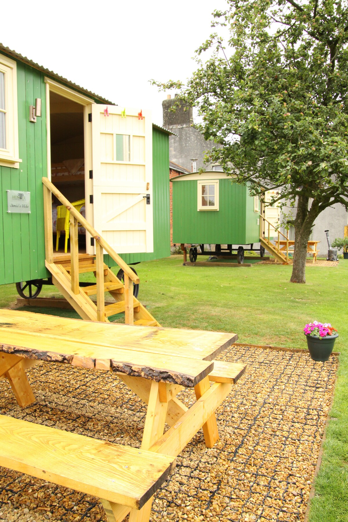 Two charming shepherd's huts are situated in a green outdoor area, surrounded by grass and trees. A wooden picnic table is placed on a gravel path in the foreground. Each hut features a set of steps leading to their entrances, creating a welcoming rustic environment.