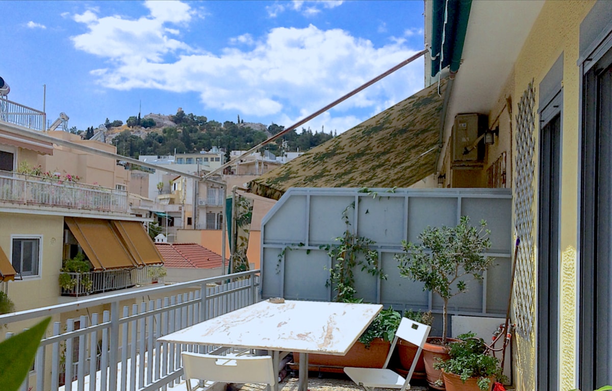 A sunny balcony space features a white table surrounded by several chairs. Potted plants are placed along the railing, adding greenery. An electronic awning provides shade, extending towards a view of Philopappos Hill and a bright blue sky scattered with clouds.