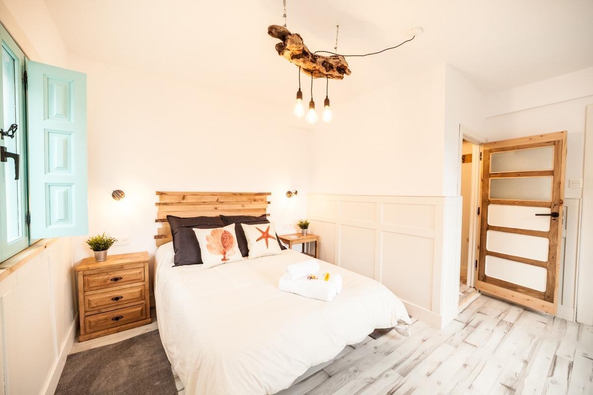 A cozy bedroom features a spacious bed adorned with decorative pillows. Natural light enters through a turquoise shuttered window. A wooden nightstand with a small plant is placed beside the bed. The ceiling is highlighted by a unique light fixture, adding character to the room.