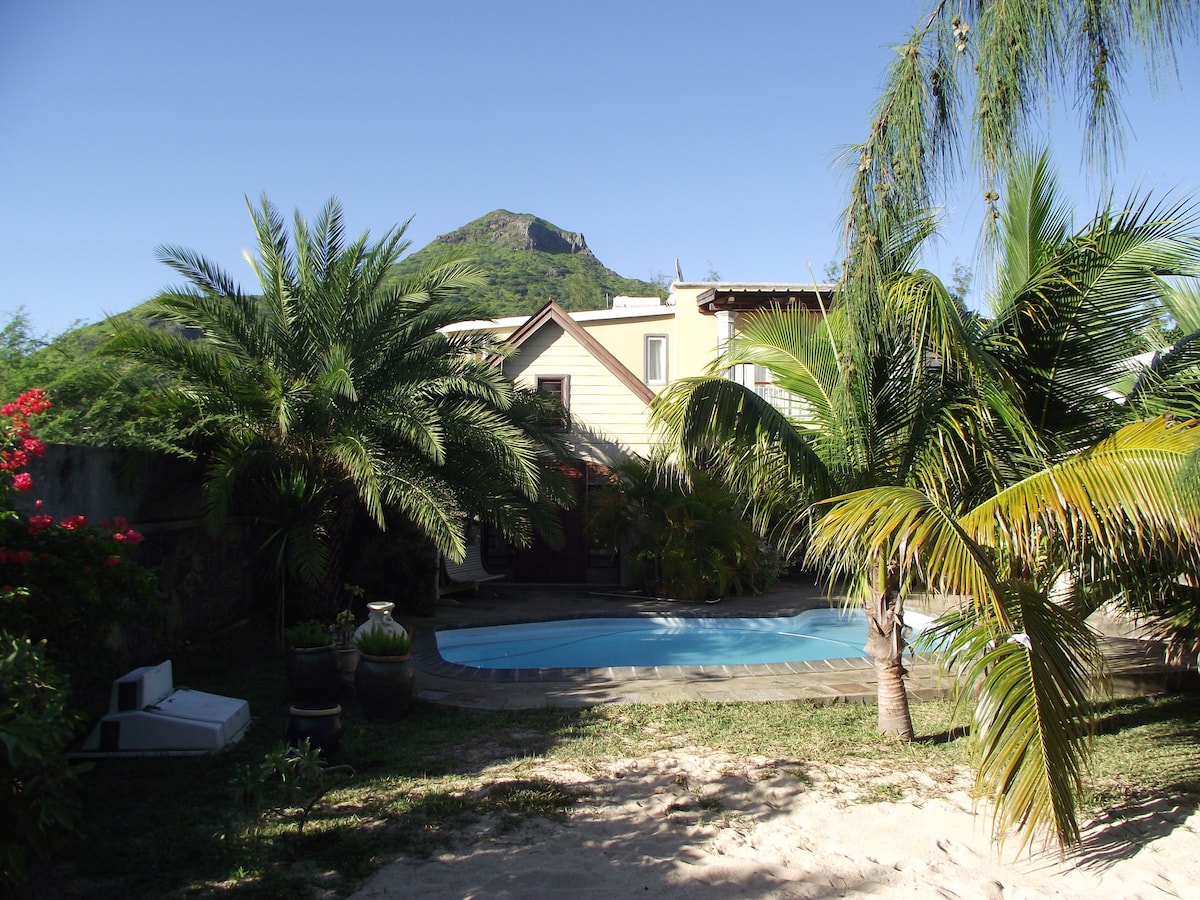 The image features a garden with lush palm trees and vibrant flowering plants. A swimming pool can be seen at the forefront, surrounded by a paved area. In the background, a two-story building is positioned against a clear blue sky, with a mountain faintly visible behind it.