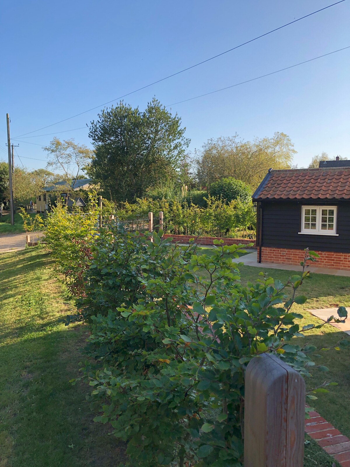 The image shows a well-maintained cottage surrounded by lush greenery and trees. A neatly trimmed hedge lines the front, with the cottage's dark exterior and red tile roof visible in the background. The area is bathed in natural light, creating a welcoming rural atmosphere.