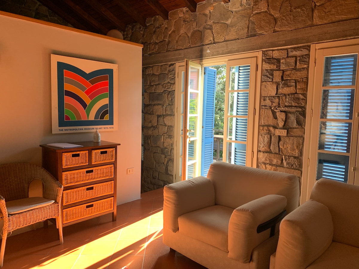 A sunlit living area features a wooden cabinet and two soft armchairs, positioned for relaxation. Natural light streams through the open French doors, complemented by stone walls and rustic wooden beams. A colorful artwork hangs on the wall, adding a vibrant touch.