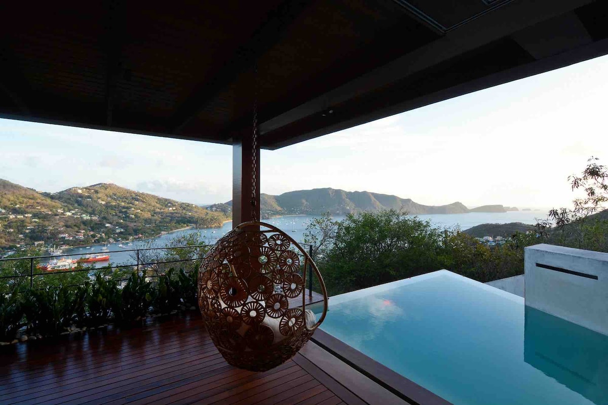 A cozy hanging chair is positioned on an outdoor deck, overlooking a serene view of rolling hills and Admiralty Bay. A private swimming pool reflects the soft colors of the sky at sunset, creating a tranquil atmosphere for relaxation.
