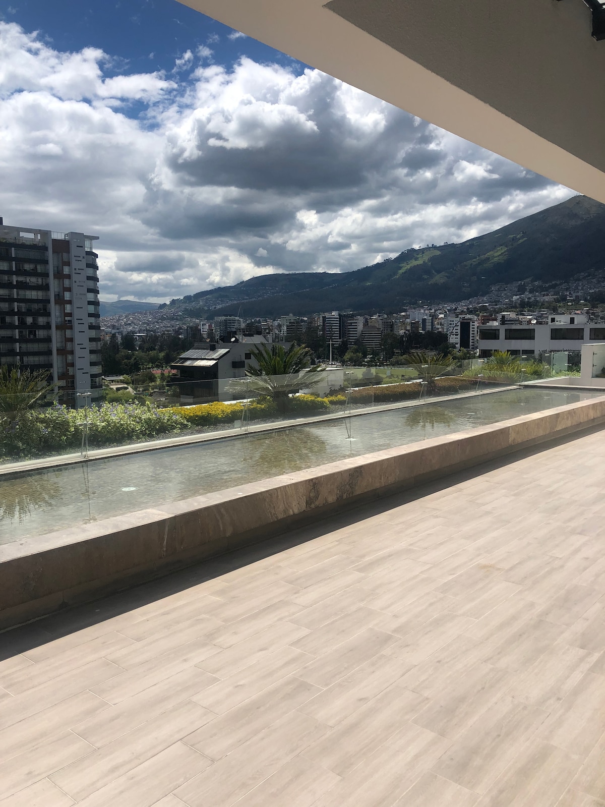 A spacious terrace offers a panoramic view of the city and mountains, framed by lush greenery. A clear glass railing ensures an unobstructed sightline over a reflective water feature, while the tiled surface provides a comfortable outdoor space.