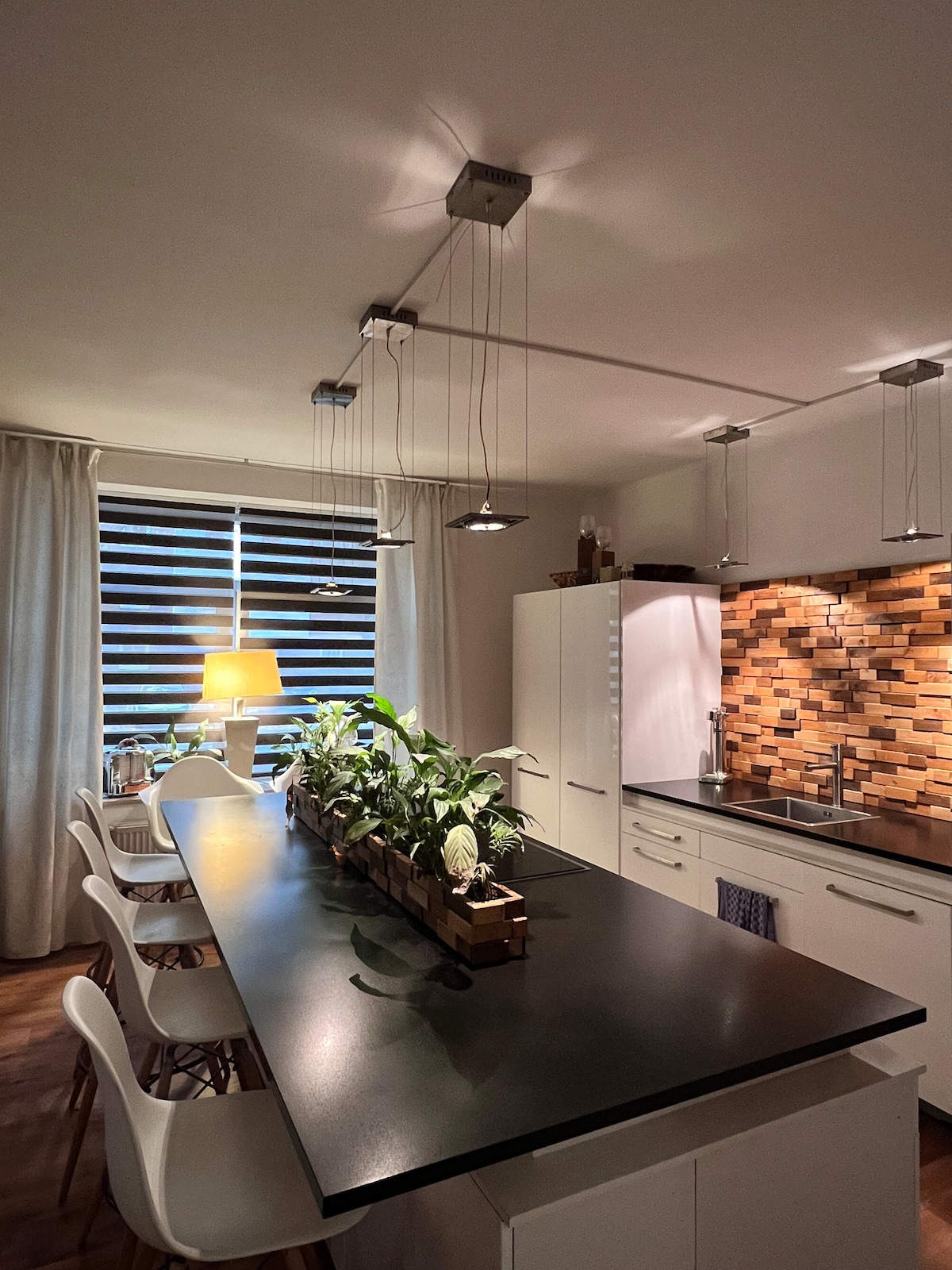 A modern kitchen is featured with an elongated dark countertop, complemented by white chairs. A row of potted plants adds greenery, while pendant lights illuminate the space above. The textured backsplash of natural wood enhances the design, and soft light filters in through the partially covered window.