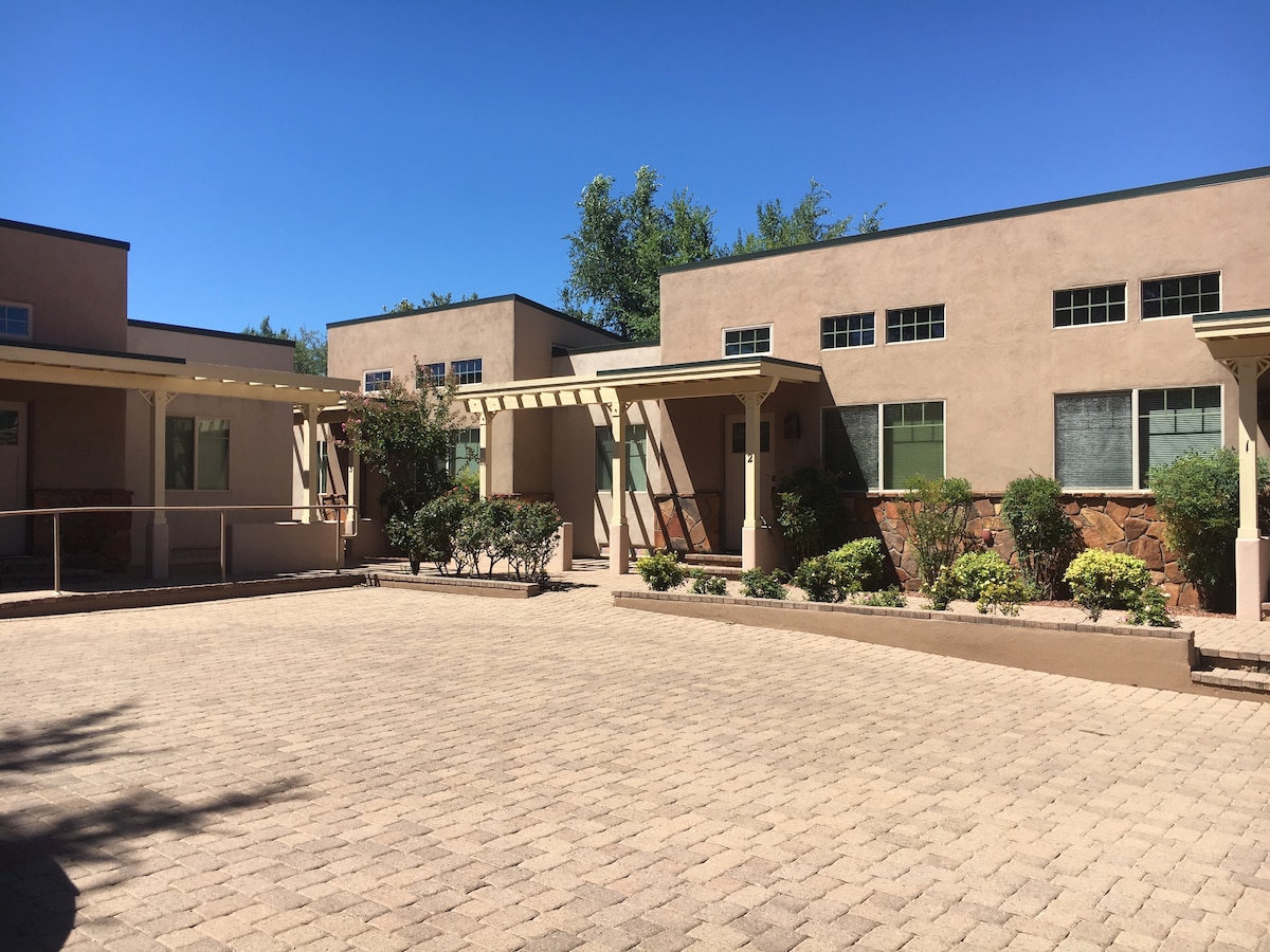 The exterior of a modern condo building is displayed, featuring a landscaped courtyard with clean, paved stones. Green bushes and small trees are strategically placed along the building's facade, complementing the architectural design. Large windows are visible, allowing for natural light.
