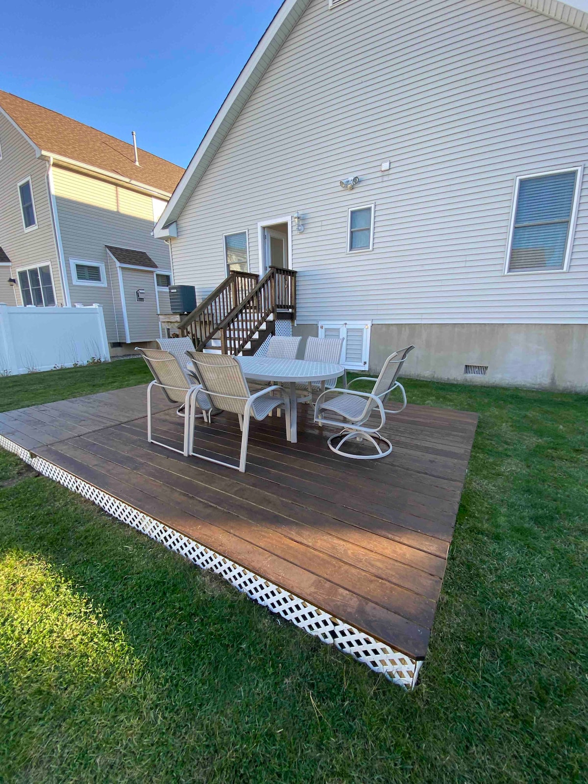 A wooden deck is shown, featuring a round table surrounded by six metal chairs. The deck is situated in the backyard of a light-colored home, with grass surrounding the area. The steps lead up to the rear entrance of the house.