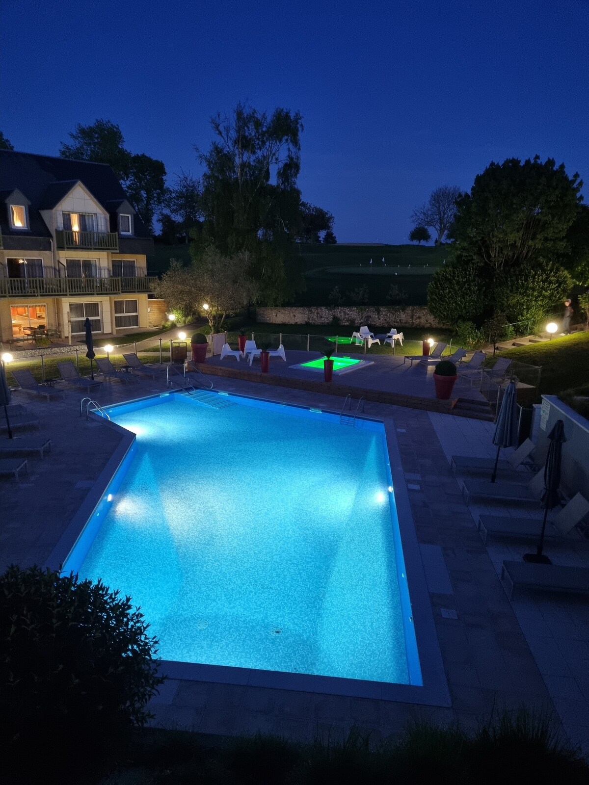 The swimming pool, illuminated in soft blue light, is surrounded by lounge chairs and shaded umbrellas. Decorative planters and landscaped greenery add to the tranquil outdoor setting. Residing nearby, a building features expansive windows that reflect the ambient lighting of the pool area.