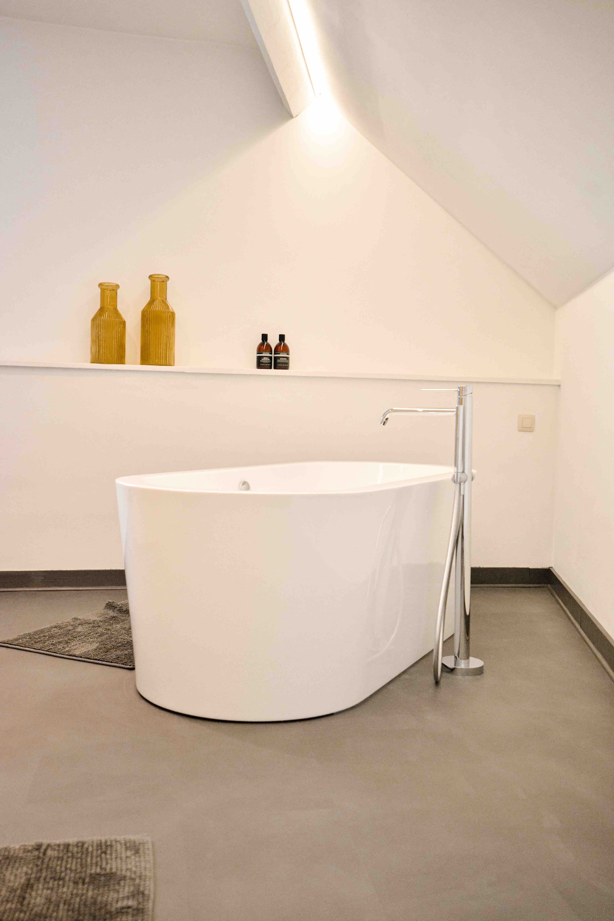 A freestanding oval bathtub is centrally placed on a smooth floor, complemented by two simple decorative vases and a couple of bottles on a shelf above. Soft lighting highlights the clean lines of the space, with a textured bath mat positioned in the foreground.