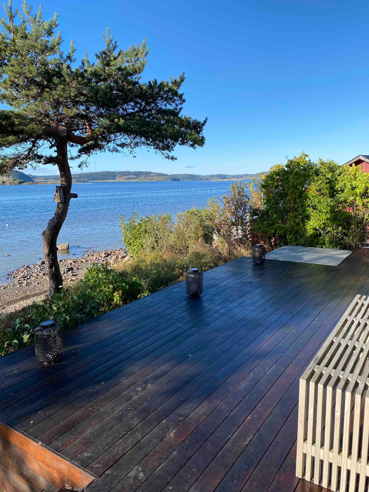 A wooden deck extends towards the water, framed by greenery and a single tree. Small lanterns are positioned along the deck, providing subtle lighting. The view showcases a calm body of water and distant hills under a clear blue sky.