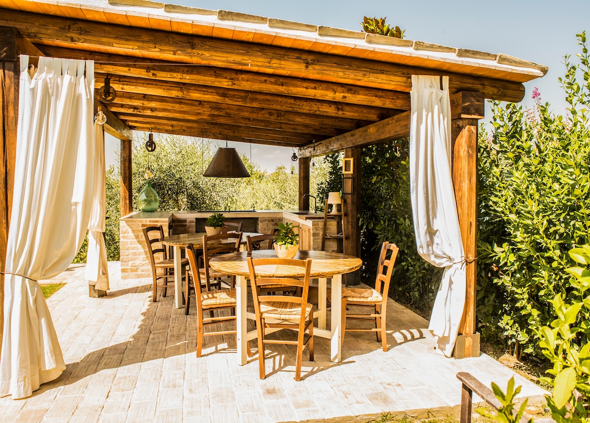 An outdoor dining area is presented under a wooden pergola, featuring a round wooden table surrounded by several wooden chairs. Sheer curtains frame the space while a built-in stone barbecue is visible in the background, complemented by lush greenery.