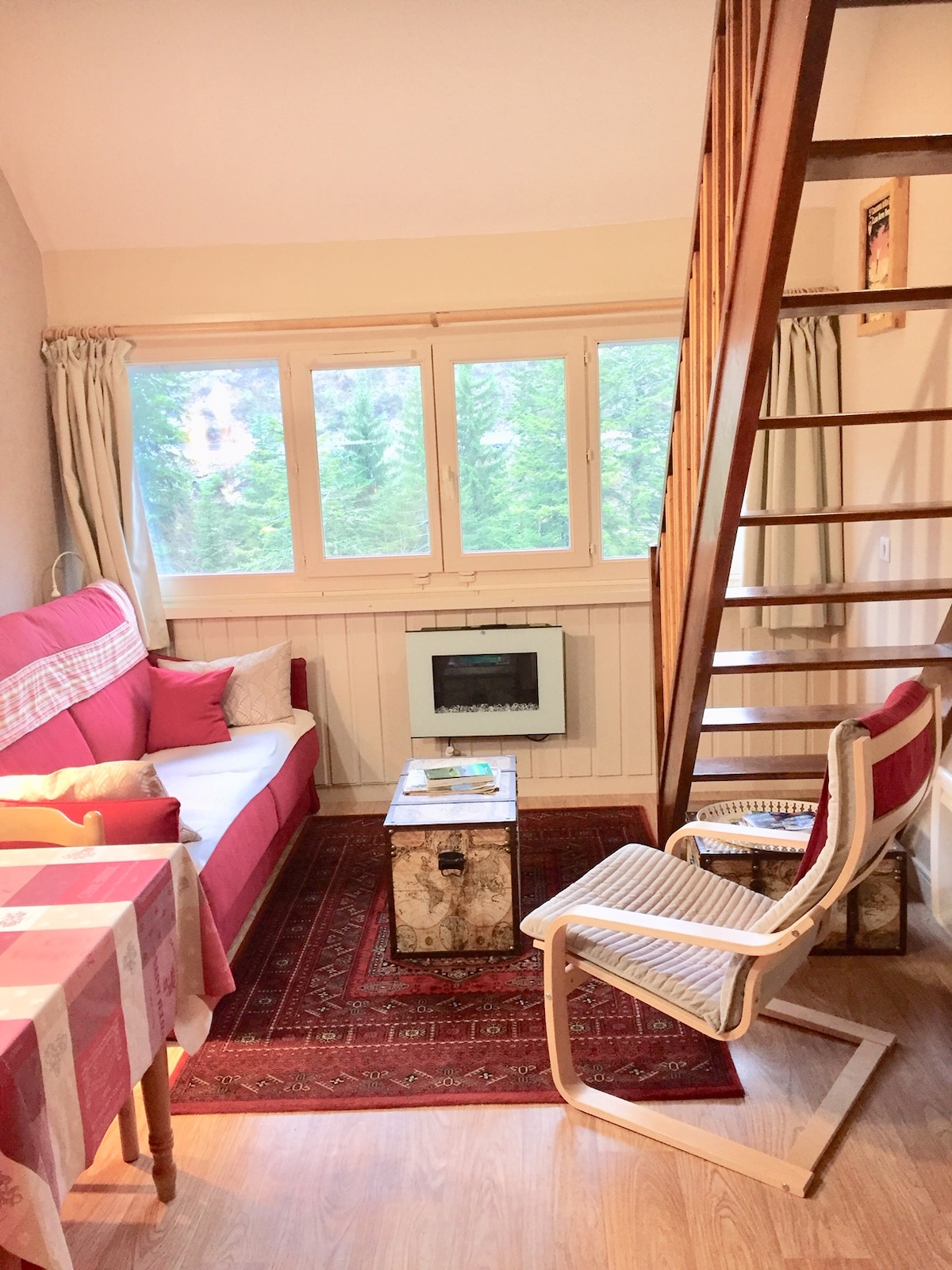 A cozy living area features a red sofa bed with decorative cushions, a rustic coffee table, and a wooden rocking chair. Large windows provide views of surrounding greenery, while a compact electric fireplace adds warmth. A staircase leads to the upper level.