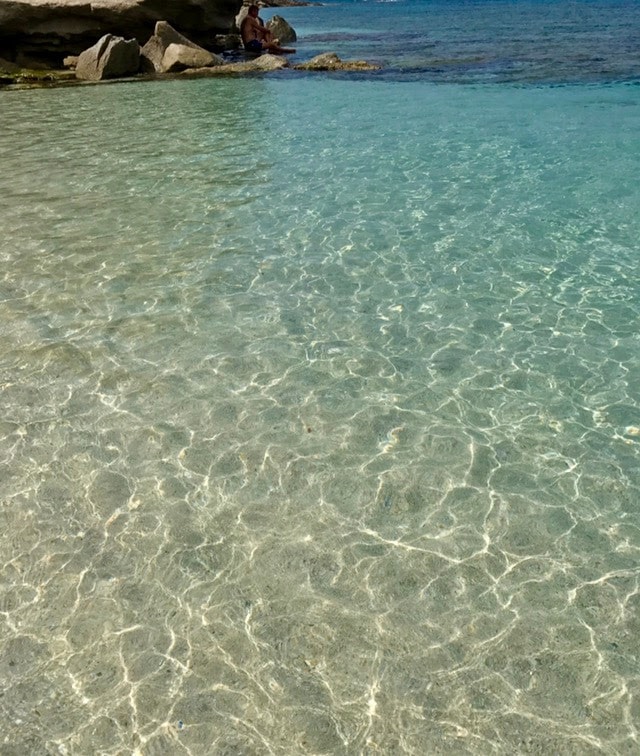 Clear turquoise water shimmers under sunlight, revealing a sandy bottom and gentle ripples. Smooth rocks are visible at the water's edge, creating a natural boundary. In the distance, a figure sits on the shore, enjoying the tranquil environment.