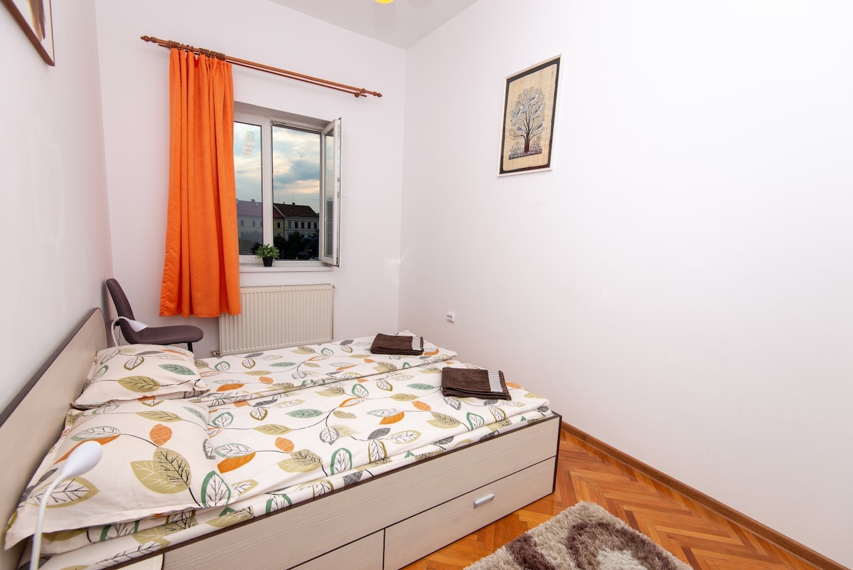 A bright bedroom features a double bed adorned with a leaf-patterned duvet. An orange curtain frames the window, allowing natural light to fill the space. A simple chair and decorative artwork add a touch of character to the room.