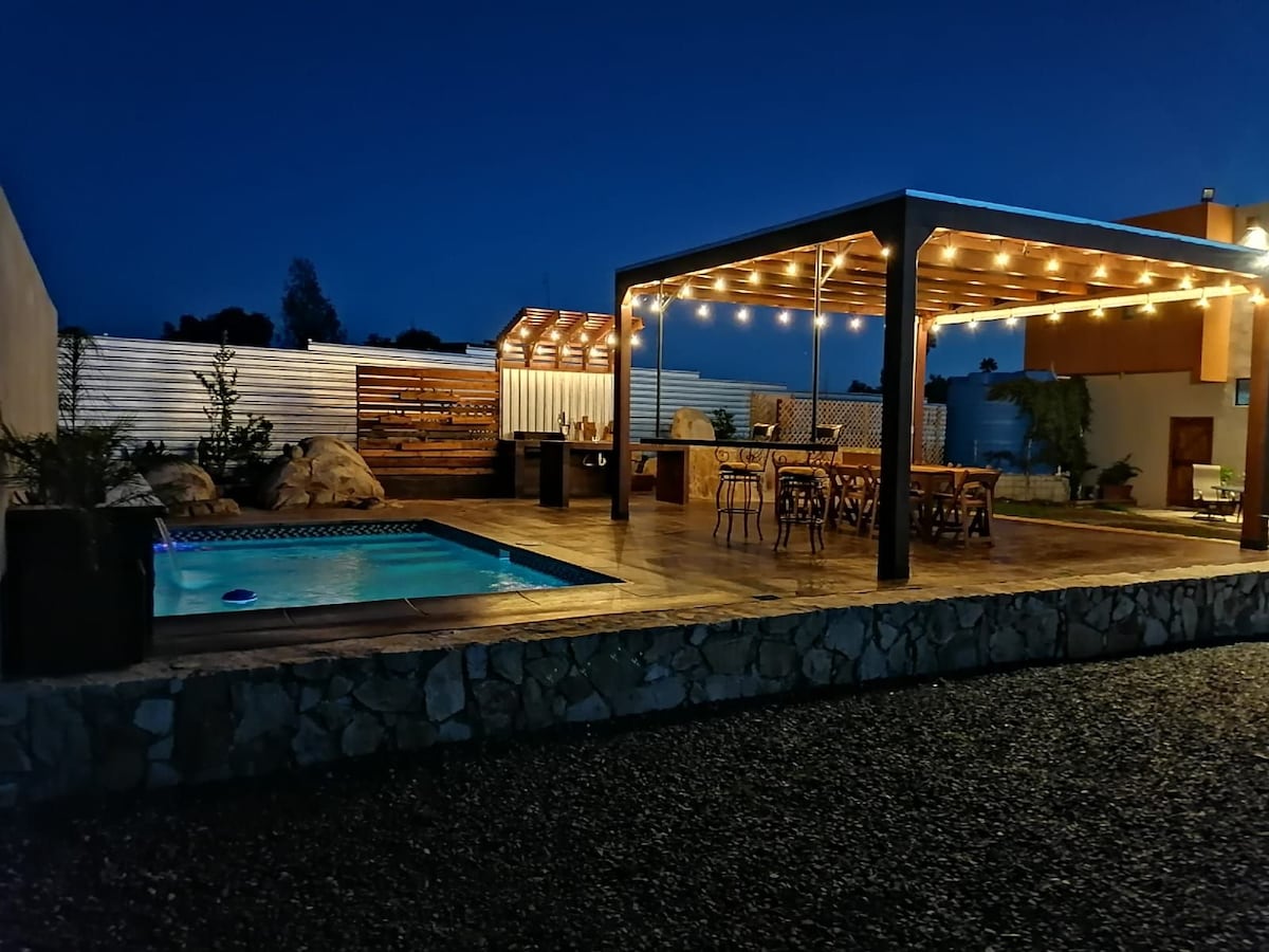An inviting outdoor area features a cozy swimming pool beneath a starlit sky. Light strands illuminate a wooden dining area with ample seating, a barbecue space, and rustic decor, creating a perfect setting for gatherings and relaxation.
