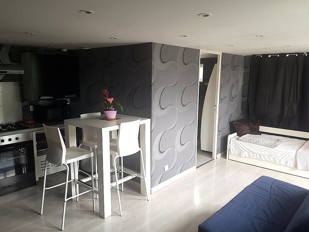An open layout captures a studio space with modern decor. A small dining area features two white bar stools beside a tall table. A comfortable sofa is positioned along one wall, while a wall-mounted television and kitchen appliances are visible in the background.