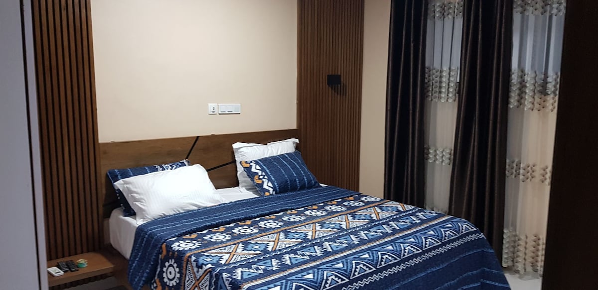 A cozy bedroom features a wooden headboard and a patterned bedspread with blue and beige tones. Two white pillows rest on the bed, while sheer curtains filter soft light from the window, creating a calm environment.