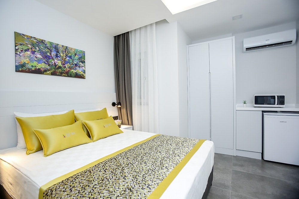 A modern bedroom features a spacious bed with yellow accent pillows and a decorative throw blanket. A vibrant artwork adorns the wall above the bed. Natural light filters through sheer curtains, illuminating a compact kitchenette with a microwave and refrigerator in the corner.