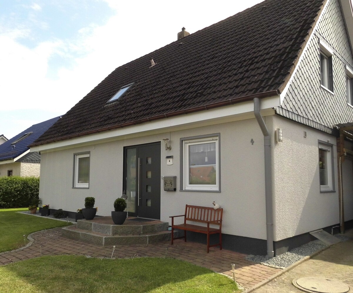 A detached two-story home is depicted, showcasing a simple exterior design. A stone pathway leads to the entrance, which features a modern door. Potted plants are arranged on either side. A small wooden bench is positioned near the entrance, surrounded by well-maintained grass.