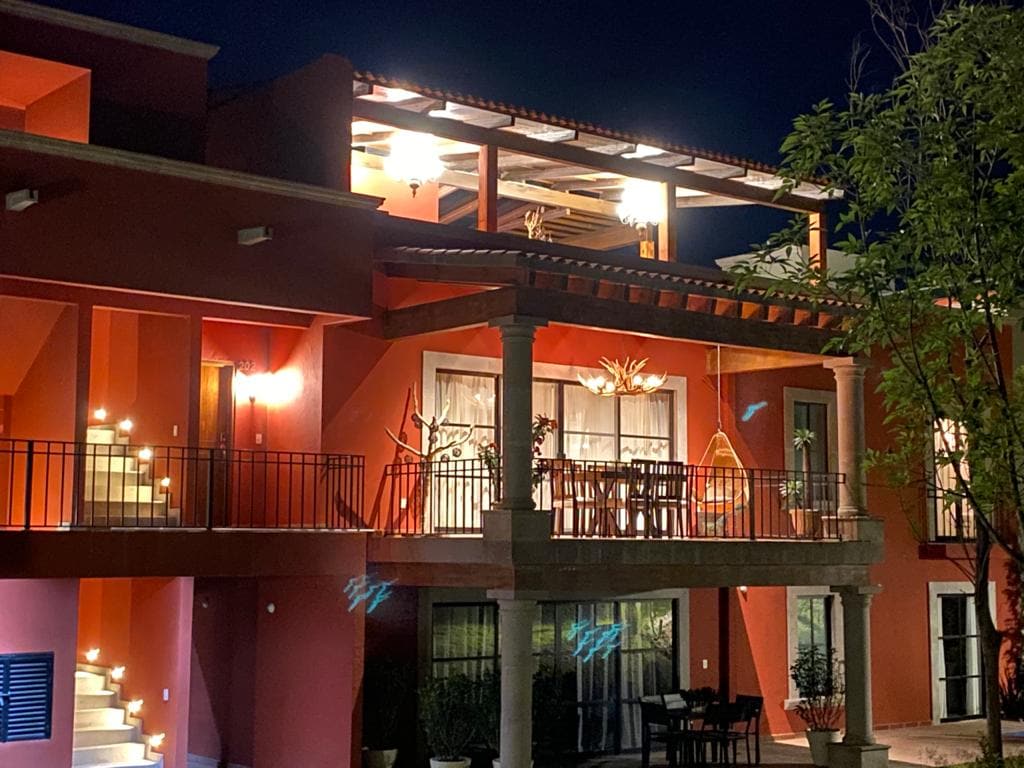 A modern penthouse is shown from an exterior viewpoint at night, featuring illuminated balconies and a spacious rooftop area. The bright lights create a welcoming ambiance against the vibrant orange facade. Dining furniture is visible on the lower terrace, surrounded by plants for added privacy.