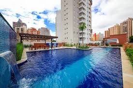 A clear blue pool is surrounded by lush greenery and modern seating areas. The pool features a waterfall element, with a backdrop of a high-rise building and a partly cloudy sky.