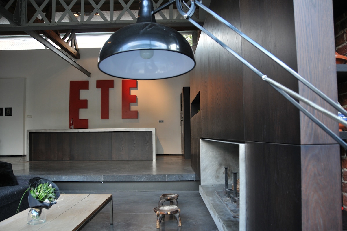 An urban loft space showcases a modern design with a large kitchen island and bold red letters spelling 'ETE' on the wall. A sleek floor lamp provides illumination, while a minimalist coffee table and a pair of small stools complete the room. A fireplace is visible in the background.