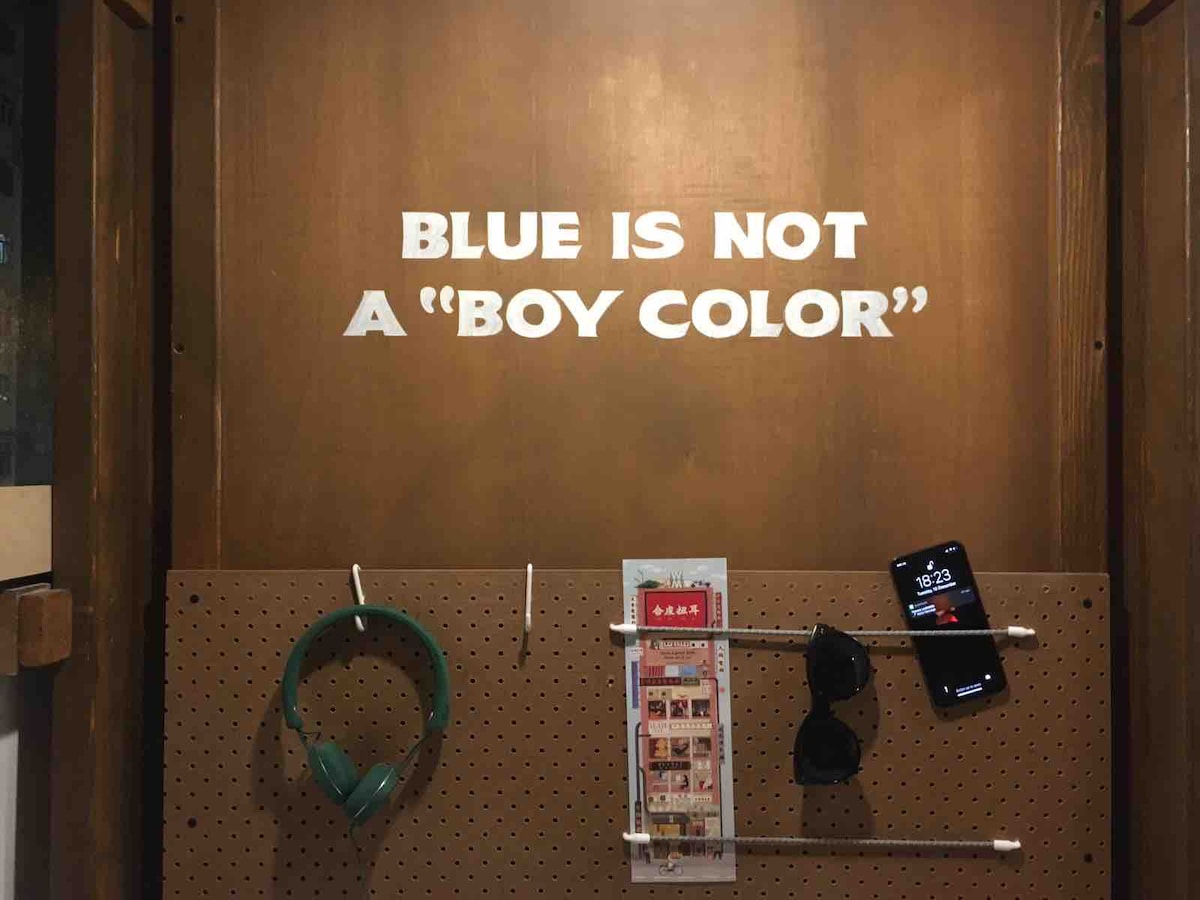 A wooden wall features a bold statement in white lettering, reading 'BLUE IS NOT A "BOY COLOR"'. Below, a green headphone rests on a peg, alongside a smartphone displaying the time, and a small brochure is placed flat for easy access.