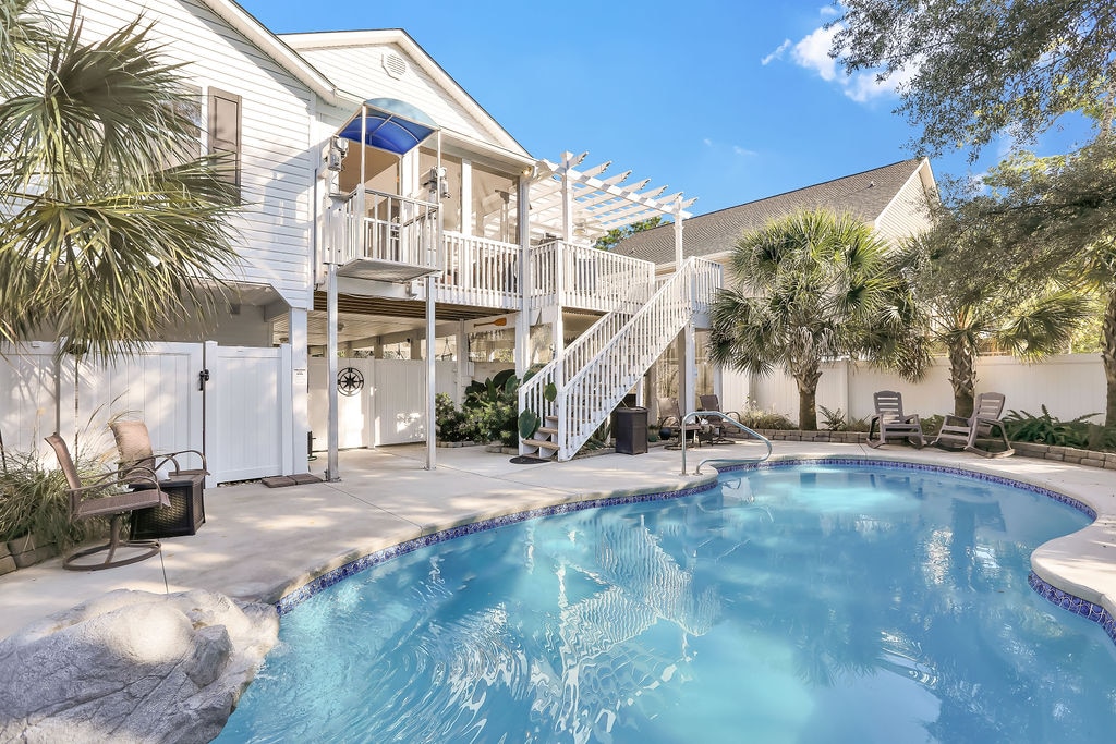 A well-maintained backyard features a heated saltwater pool with a gently curved edge. Comfortable lounge chairs are arranged around the pool, with a backdrop of palm trees and lush greenery. A spacious deck is visible above, providing shaded seating areas.
