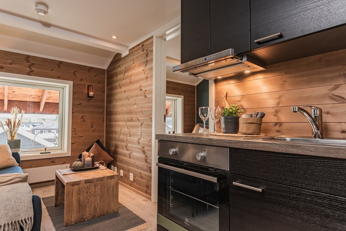 The image shows a well-appointed kitchenette featuring dark cabinetry, an oven, and a stovetop. A wooden table is positioned nearby, accompanied by decorative items. Large windows provide ample natural light, illuminating the warm wood paneling that adds a cozy touch to the living area.
