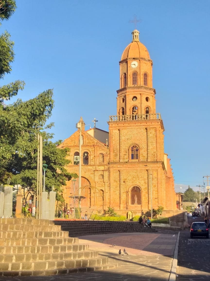 A historical brick church with a prominent clock tower is displayed against a clear blue sky. Green trees frame the structure, while the surrounding area features cobblestone paths and a hint of local activity in the background.