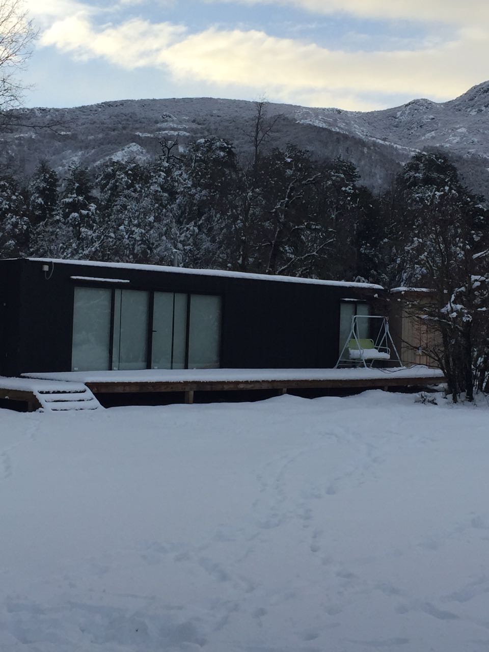 A modern cabin is positioned amidst a snowy landscape, surrounded by trees and mountains. Large glass doors provide unobstructed views of the serene scenery, and a wooden deck extends outward, inviting outdoor relaxation in a tranquil setting.